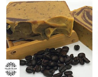 May include: Two bars of coffee-scented soap with brown and beige swirls, sitting on a wooden soap dish. Coffee beans are scattered around the soap dish.