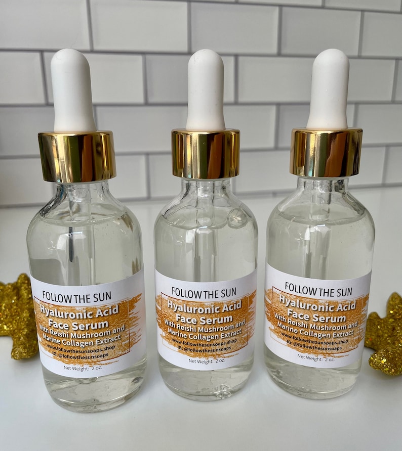 May include: Three glass bottles with gold lids and droppers filled with clear liquid. Each bottle has a white label with the text "FOLLOW THE SUN" and "Hyaluronic Acid Face Serum" with Reishi Mushroom and Marine Collagen Extract. The bottles are on a white surface with gold stars.