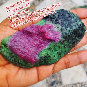 May include: A raw ruby zoisite specimen featuring a vibrant magenta-pink center surrounded by mottled green and black mineral inclusions. The stone is labeled with the text A) 90X50X25MM, 1120 CARATS, 224 GRAMS, RARE GENUINE UNIQUE HUGE EXCELLENT RUBY ZOSITE.