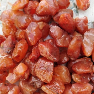 Puede incluir: Una colección de piedras de cornalina en bruto y pulidas en varios tonos de naranja, naranja rojizo y ámbar intenso. Las piedras tienen formas orgánicas irregulares con superficies lisas y pulidas que exhiben una calidad translúcida.