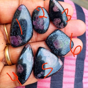 May include: Six polished cabochon gemstones featuring a black base with deep magenta and pink inclusions. Each stone is numbered in red ink from one to six, displaying various shapes including teardrop, oval, marquise, and triangular forms.