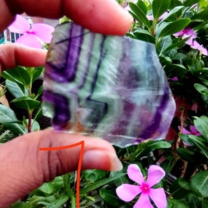 Puede incluir: Un trozo plano y transl&uacute;cido de mineral de fluorita que presenta bandas angulares distintivas de color p&uacute;rpura intenso y verde p&aacute;lido. La piedra tiene un borde irregular y dentado, mostrando un patr&oacute;n de color natural y estratificado.