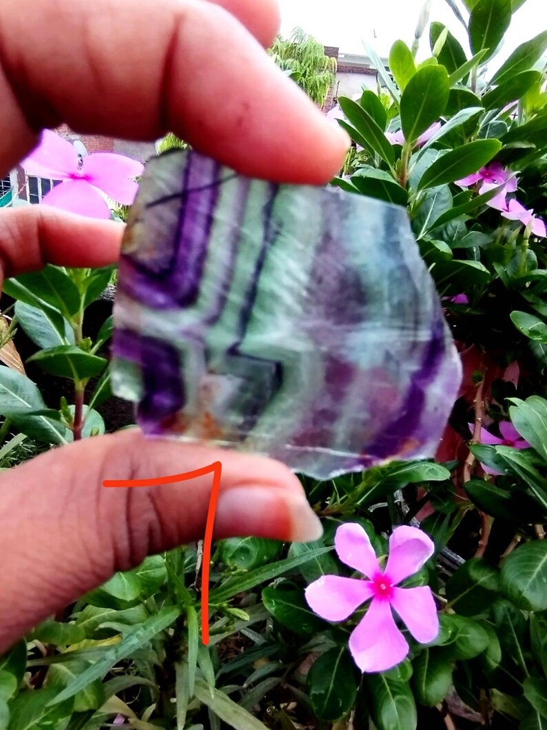 Puede incluir: Un trozo plano y transl&uacute;cido de mineral de fluorita que presenta bandas angulares distintivas de color p&uacute;rpura intenso y verde p&aacute;lido. La piedra tiene un borde irregular y dentado, mostrando un patr&oacute;n de color natural y estratificado.