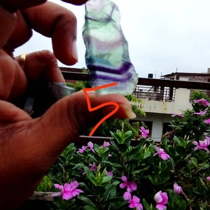 Puede incluir: Un esp&eacute;cimen de cristal de fluorita crudo y transl&uacute;cido que presenta bandas horizontales distintivas de capas minerales de color p&uacute;rpura, verde y transparente. La piedra tiene una forma irregular y dentada con una textura superficial lisa.