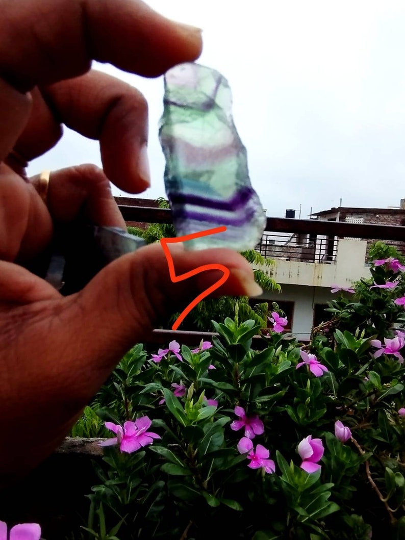 Puede incluir: Un esp&eacute;cimen de cristal de fluorita crudo y transl&uacute;cido que presenta bandas horizontales distintivas de capas minerales de color p&uacute;rpura, verde y transparente. La piedra tiene una forma irregular y dentada con una textura superficial lisa.