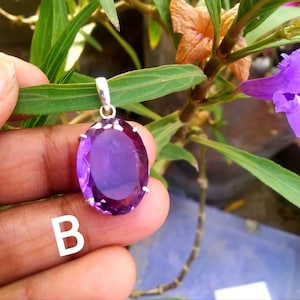 May include: An oval-shaped purple amethyst gemstone pendant set in a silver-toned metal prong setting with a bail at the top. The letter B is printed in white on the lower left side of the image, which features a faceted, translucent stone.