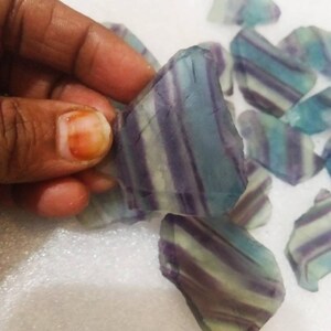 May include: A collection of raw fluorite mineral specimens featuring distinct, parallel bands of translucent purple, teal, and pale green colors. Each piece has an irregular, angular shape with a smooth, glass-like texture and visible internal layering.