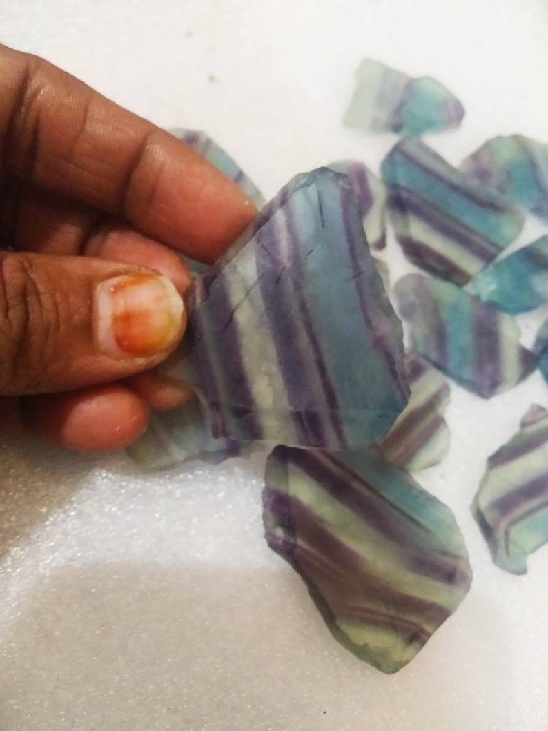 May include: A collection of raw fluorite mineral specimens featuring distinct, parallel bands of translucent purple, teal, and pale green colors. Each piece has an irregular, angular shape with a smooth, glass-like texture and visible internal layering.