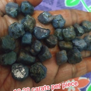 Puede incluir: Una colección de piedras preciosas de zafiro azul en bruto y sin pulir, con formas irregulares y variados tonos azul oscuro. Cada piedra individual pesa entre 10 y 20 quilates. La superficie de las piedras muestra texturas, inclusiones y facetas naturales.