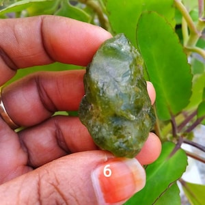 Puede incluir: Una piedra preciosa en bruto, translúcida y de color verde oliva con una textura superficial irregular y multifacética. La piedra muestra tonos verde bosque profundo y formaciones cristalinas naturales en toda su estructura, marcada con el número 6 en blanco.