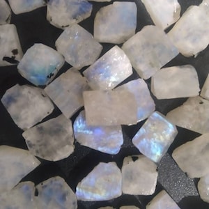 Puede incluir: Una colección de pequeñas piezas rectangulares y translúcidas de piedra lunar con una apariencia blanca lechosa y destellos azules iridiscentes. Cada piedra tiene una superficie lisa y pulida con bordes irregulares, mostrando diversos grados de reflexión de luz interna.
