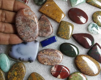 May include: A collection of polished, multi-colored cabochon gemstones in various shapes and sizes, including oval, teardrop, and rectangular forms. The stones feature diverse patterns and textures in shades of red, green, brown, blue, and white.