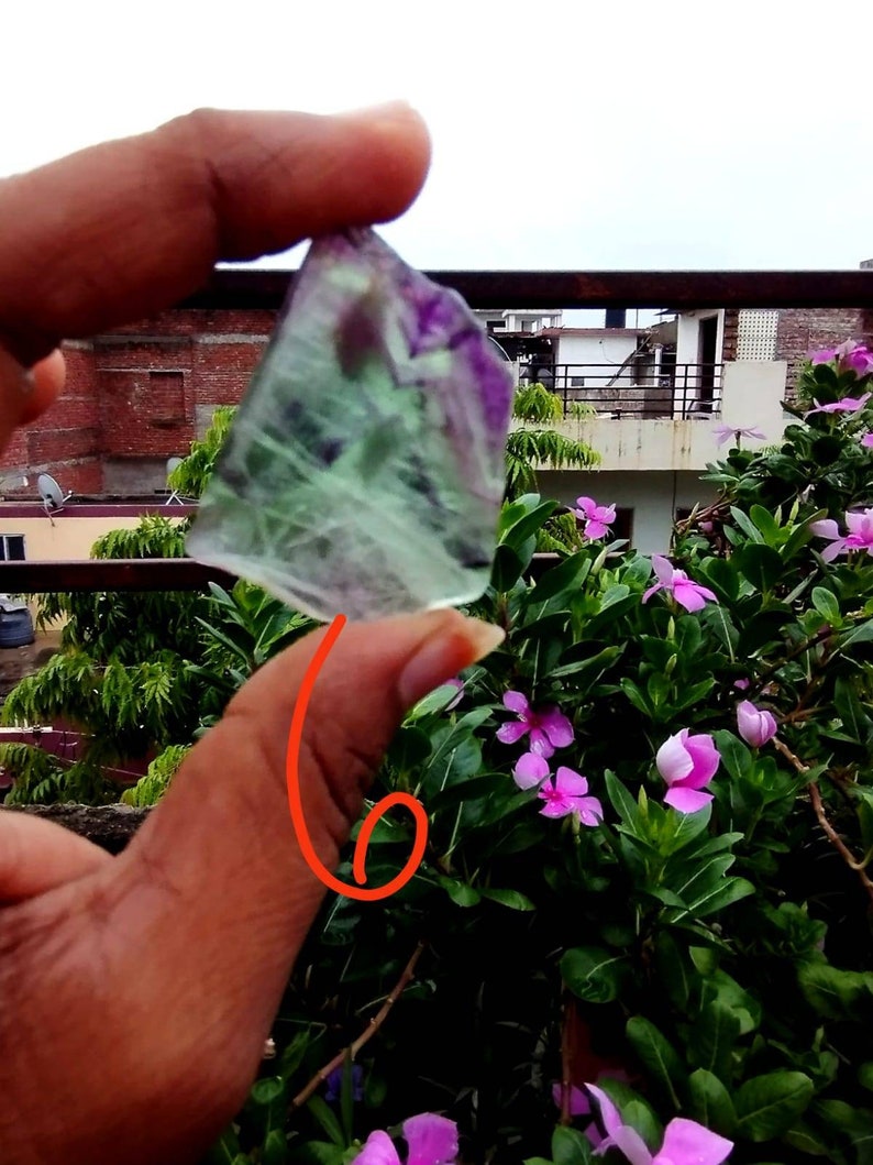 Puede incluir: Un trozo de cristal de fluorita transl&uacute;cido de forma irregular que presenta bandas distintivas de color verde claro y morado intenso. El esp&eacute;cimen mineral muestra una superficie lisa y pulida con fracturas internas visibles y zonificaci&oacute;n.