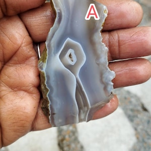 May include: A translucent grey and white agate slab featuring a central crystalline cavity and natural banded patterns, marked with the letter A in white. The stone has irregular, rough, earth-toned edges and a smooth, polished surface texture.