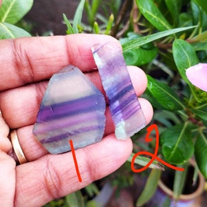 Puede incluir: Dos piezas planas y pulidas de cristal de fluorita bandeada. La pieza m&aacute;s grande tiene forma pentagonal, mientras que la m&aacute;s peque&ntilde;a es rectangular. Ambas piedras presentan capas transl&uacute;cidas de color p&uacute;rpura intenso, violeta claro y verde p&aacute;lido.