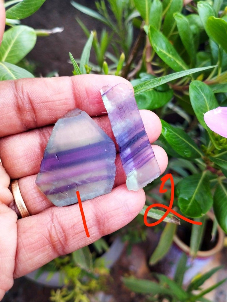 Puede incluir: Dos piezas planas y pulidas de cristal de fluorita bandeada. La pieza m&aacute;s grande tiene forma pentagonal, mientras que la m&aacute;s peque&ntilde;a es rectangular. Ambas piedras presentan capas transl&uacute;cidas de color p&uacute;rpura intenso, violeta claro y verde p&aacute;lido.