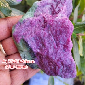 May include: A raw, unpolished mineral specimen featuring a vibrant magenta-pink crystalline section contrasted against a mottled green base. The stone is labeled with the dimensions 75x50x35mm and a weight of 1300 carats, clearly printed on the surface.