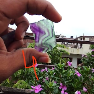 Puede incluir: Un trozo de fluorita transl&uacute;cida de forma irregular con bandas distintivas de color p&uacute;rpura y verde claro. La piedra muestra un patr&oacute;n geom&eacute;trico natural dentro de su estructura, sostenida contra un cielo brillante y nublado.