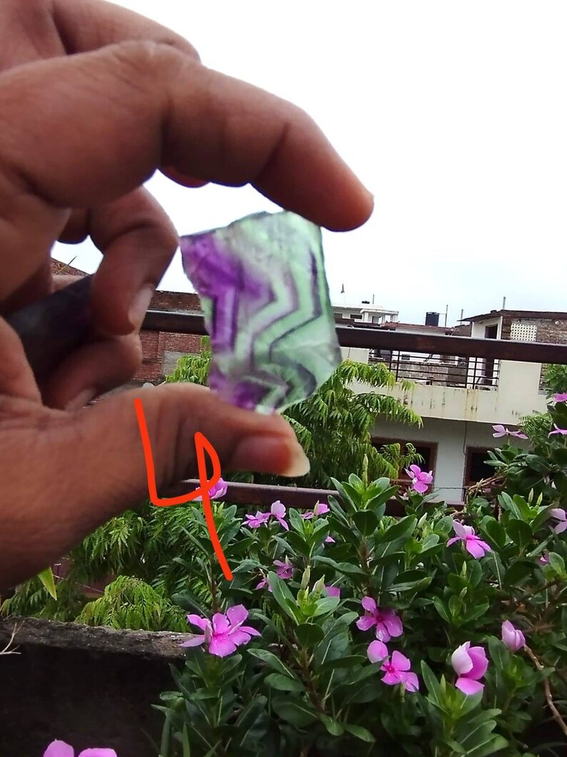 Puede incluir: Un trozo de fluorita transl&uacute;cida de forma irregular con bandas distintivas de color p&uacute;rpura y verde claro. La piedra muestra un patr&oacute;n geom&eacute;trico natural dentro de su estructura, sostenida contra un cielo brillante y nublado.