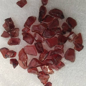 May include: A collection of small, irregular, deep red garnet gemstones with a polished, translucent appearance. Each stone features unique, angular facets and varying shapes, displaying a rich, dark crimson hue throughout the entire group.
