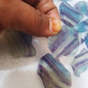 May include: A collection of raw fluorite mineral specimens featuring distinct banded patterns in shades of translucent purple, blue, and pale green. Each piece displays a smooth, polished surface with visible internal layers and irregular edges.