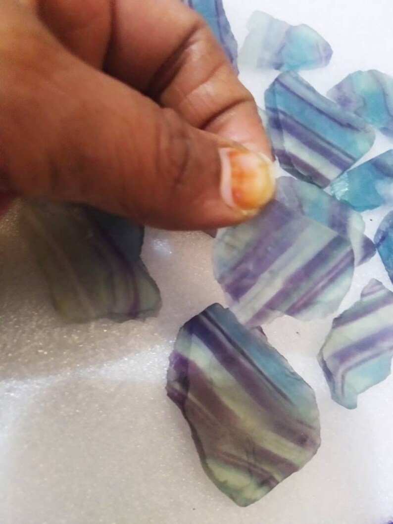 May include: A collection of raw fluorite mineral specimens featuring distinct banded patterns in shades of translucent purple, blue, and pale green. Each piece displays a smooth, polished surface with visible internal layers and irregular edges.