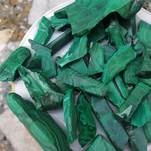 Puede incluir: Una colección de fragmentos de piedra de malaquita en bruto con patrones de bandas en verde bosque profundo y verde claro. Las piezas presentan formas naturales e irregulares con superficies lisas y pulidas que muestran estrías circulares.