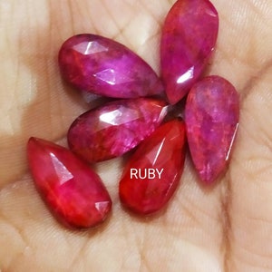 May include: Five faceted, pear-shaped ruby gemstones with deep red and magenta hues. The stones feature a polished, reflective surface with visible internal inclusions. The word RUBY is printed in white text between the center stones.