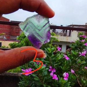 Puede incluir: Un esp&eacute;cimen mineral angular y transl&uacute;cido que presenta bandas distintivas de color p&uacute;rpura y verde. El cristal muestra un patr&oacute;n geom&eacute;trico en capas en toda su estructura, con un borde irregular en la parte superior y una base puntiaguda.