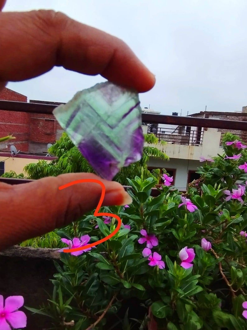 Puede incluir: Un esp&eacute;cimen mineral angular y transl&uacute;cido que presenta bandas distintivas de color p&uacute;rpura y verde. El cristal muestra un patr&oacute;n geom&eacute;trico en capas en toda su estructura, con un borde irregular en la parte superior y una base puntiaguda.