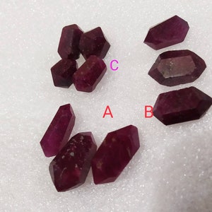 May include: A collection of deep red, double-terminated ruby crystals arranged in groups. The stones are labeled with the letters A, B, and C in red and pink text. Each crystal features a hexagonal, elongated shape with pointed ends and a faceted surface.