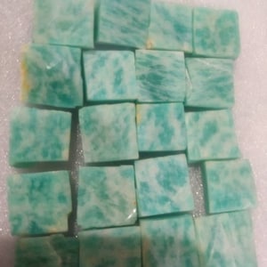 以下が含まれることがあります： グリッド状に配置された17個の正方形にカットされたアマゾナイトの石のコレクション。各石は、不透明なターコイズブルーとティールブルーの色合いに、白い脈模様と時折見られるかすかな黄色の鉱物の内包物が混ざり合ったユニークなパターンを表面全体に示しています。
