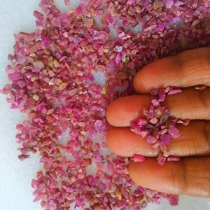 May include: A collection of small, raw, unpolished ruby gemstone fragments. The stones are vibrant pinkish-red in color, featuring irregular, jagged shapes and varying textures. These loose, granular mineral pieces are scattered across a surface.