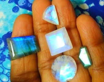 May include: A collection of five faceted gemstones including a rectangular blue labradorite, a triangular moonstone, a square moonstone, a round moonstone, and a small elongated trapezoidal green gemstone, all displaying iridescent light reflections.