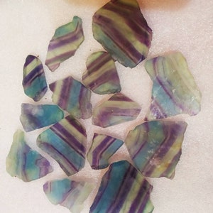 May include: A collection of twelve raw fluorite mineral specimens featuring distinct banded patterns in shades of purple, blue, and pale yellow. Each piece displays a translucent, crystalline structure with irregular, jagged edges and smooth surfaces.