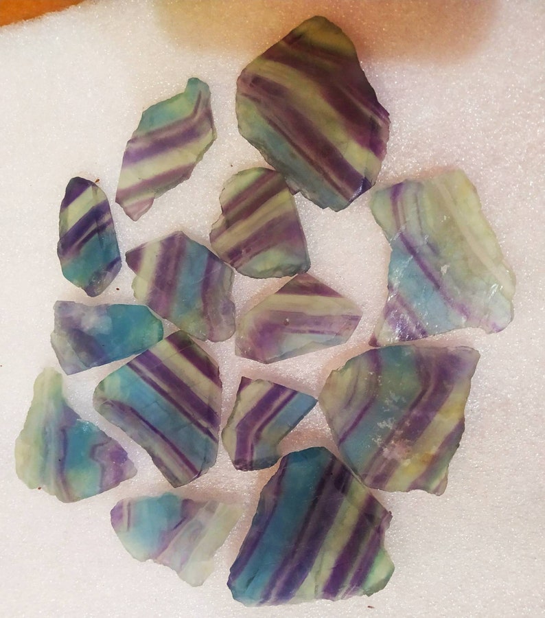 May include: A collection of twelve raw fluorite mineral specimens featuring distinct banded patterns in shades of purple, blue, and pale yellow. Each piece displays a translucent, crystalline structure with irregular, jagged edges and smooth surfaces.