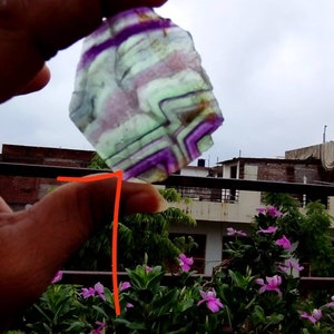 Puede incluir: Una rebanada transl&uacute;cida de fluorita de forma irregular con bandas angulares distintivas de color p&uacute;rpura, verde y blanco. La piedra muestra un patr&oacute;n geom&eacute;trico natural y estratificado en toda su estructura cristalina.