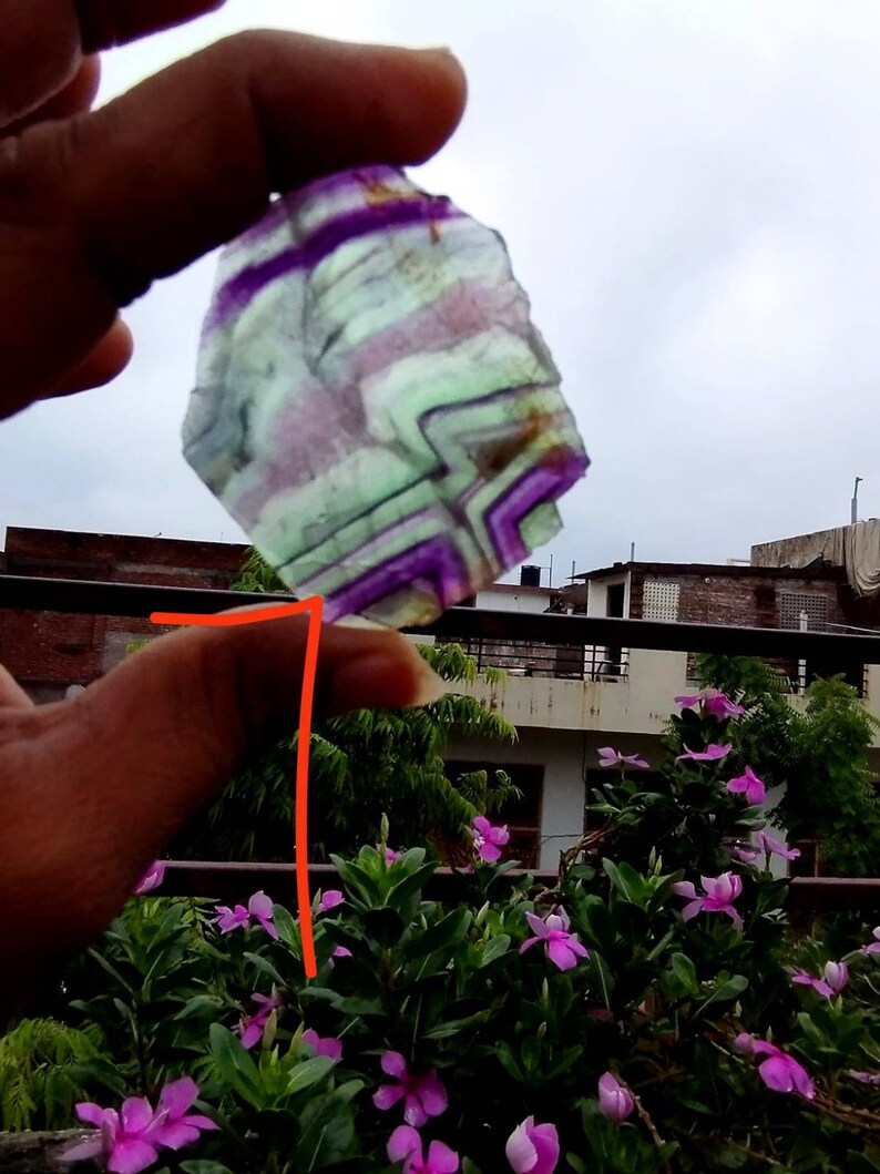 Puede incluir: Una rebanada transl&uacute;cida de fluorita de forma irregular con bandas angulares distintivas de color p&uacute;rpura, verde y blanco. La piedra muestra un patr&oacute;n geom&eacute;trico natural y estratificado en toda su estructura cristalina.
