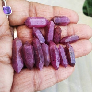 May include: A collection of eleven small, double-terminated ruby crystals with a deep magenta-red hue and natural faceted surfaces. Each stone features a pointed end on both sides, showcasing a raw, semi-translucent texture and varied color depth.