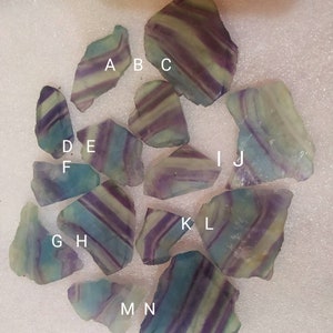 May include: A collection of fourteen raw fluorite mineral specimens, each featuring distinct banded patterns of purple, green, and translucent white. The stones are labeled with white letters A through N, showcasing natural jagged edges and varied shapes.