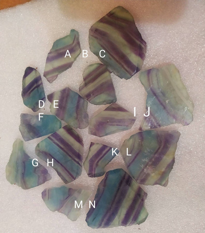 May include: A collection of fourteen raw fluorite mineral specimens, each featuring distinct banded patterns of purple, green, and translucent white. The stones are labeled with white letters A through N, showcasing natural jagged edges and varied shapes.