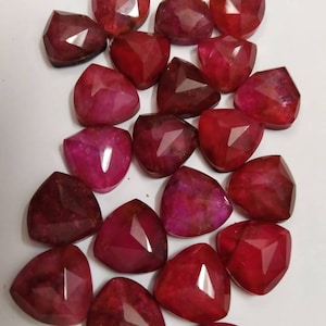 Puede incluir: Una colección de piedras preciosas facetadas sueltas de color rojo intenso. Cada piedra presenta una forma triangular con múltiples superficies planas y pulidas que reflejan la luz. Las piedras varían ligeramente en su tono de rojo, desde carmesí oscuro hasta magenta brillante, y están dispuestas en un patrón disperso.