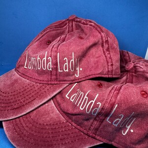 May include: Two maroon baseball caps with a distressed, washed-out finish. Each cap features the text "Lambda Lady." embroidered in white, sans-serif lettering across the front panel. The caps have a curved brim and a structured crown design.