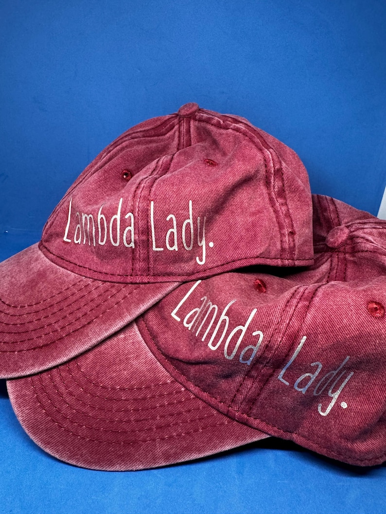 May include: Two maroon baseball caps with a distressed, washed-out finish. Each cap features the text "Lambda Lady." embroidered in white, sans-serif lettering across the front panel. The caps have a curved brim and a structured crown design.