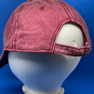May include: A faded red baseball cap with a six-panel construction and a stitched eyelet on the crown. The back features an adjustable fabric strap with a metallic buckle closure. The cap is displayed on a white mannequin head with a pearl necklace.