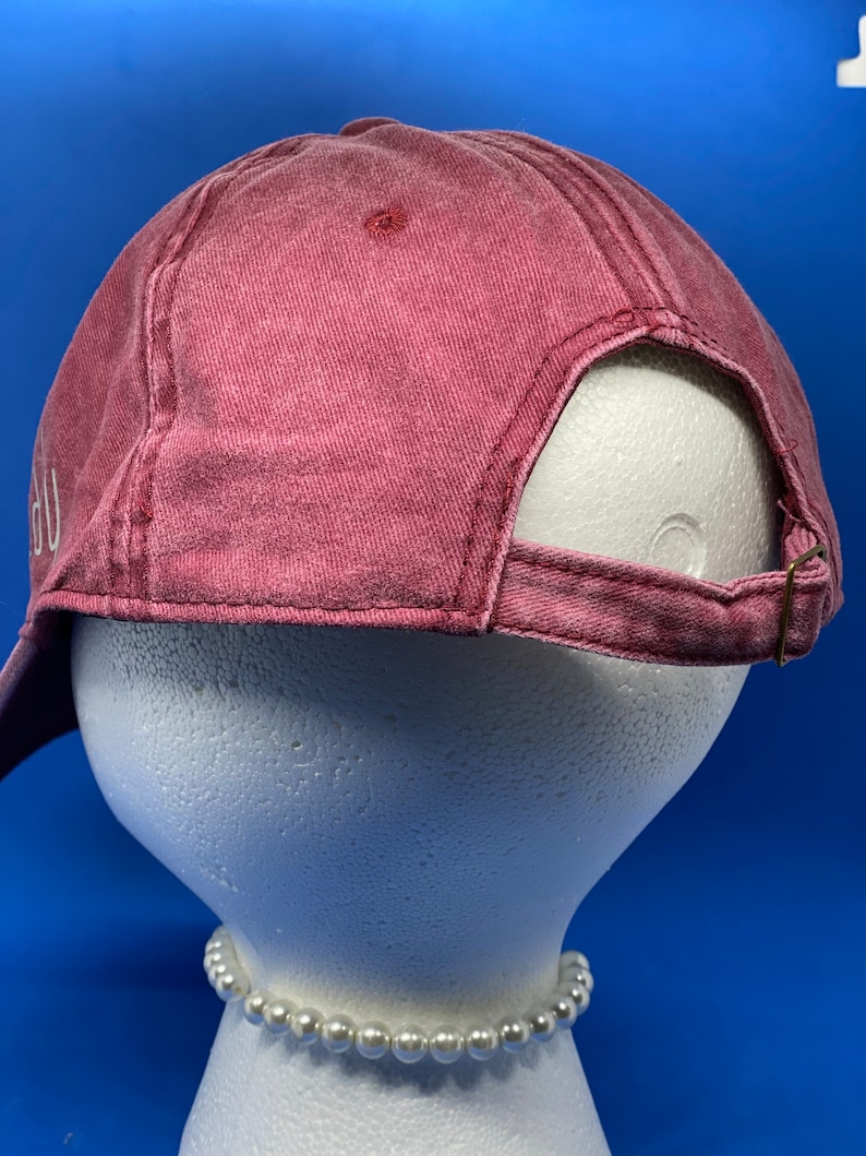 May include: A faded red baseball cap with a six-panel construction and a stitched eyelet on the crown. The back features an adjustable fabric strap with a metallic buckle closure. The cap is displayed on a white mannequin head with a pearl necklace.