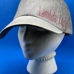 May include: A grey baseball cap with a distressed fabric texture featuring the text Lambda Lady in red lettering on the front panel. The cap has a curved brim and a structured crown, displayed on a white mannequin head against a blue background.