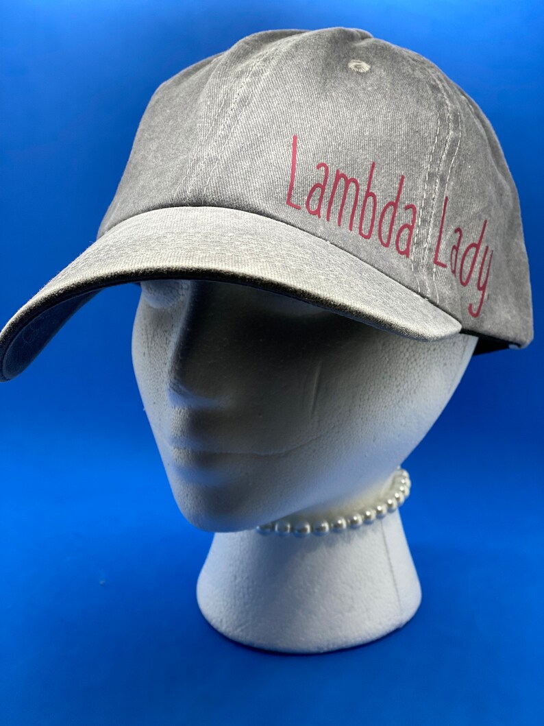 May include: A grey baseball cap with a distressed fabric texture featuring the text Lambda Lady in red lettering on the front panel. The cap has a curved brim and a structured crown, displayed on a white mannequin head against a blue background.