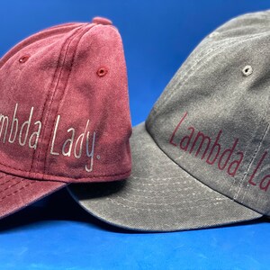 May include: Two baseball caps, one burgundy and one gray, both with the text "Lambda Lady" embroidered on them. The burgundy cap has silver embroidery, and the gray cap has burgundy embroidery.