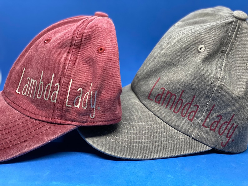 May include: Two baseball caps with a distressed finish. The left cap is maroon with silver text that reads Lambda Lady. The right cap is charcoal gray with dark red text that reads Lambda Lady. Both caps feature stitched eyelets and curved brims.